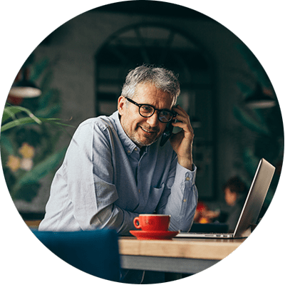 grey haired man wearing glasses sat at a table on a mobile phone working on a laptop